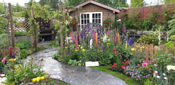 A stone grey S curved pathway leads through garden beds filled with many flowers. There is a wooden bench next to a shed.